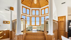 A spacious interior with a high ceiling, large windows, and a central staircase with wooden railings. - Sea Spirit OBX Dog Friendly Oceanfront HotTub (Corolla)
