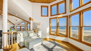 A spacious living room with a large bay window overlooking a beach, wooden flooring, and a cozy seating area. - Sea Spirit OBX Dog Friendly Oceanfront HotTub (Corolla)