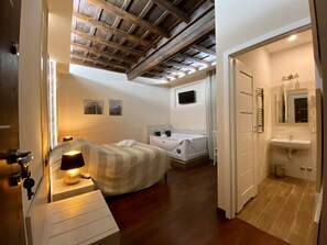 A bedroom with a bed, bedside lamp, wooden ceiling beams, a TV, and a bathroom with a sink and mirror. - Campo Marzio Hotelier (Rome)