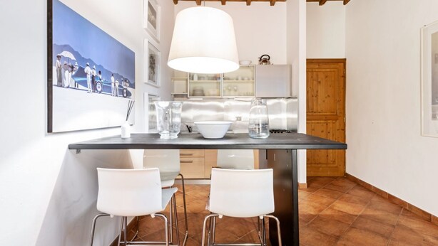 Una cucina con angolo bar, sedie bianche, una lampada a sospensione e un quadro appeso al muro.