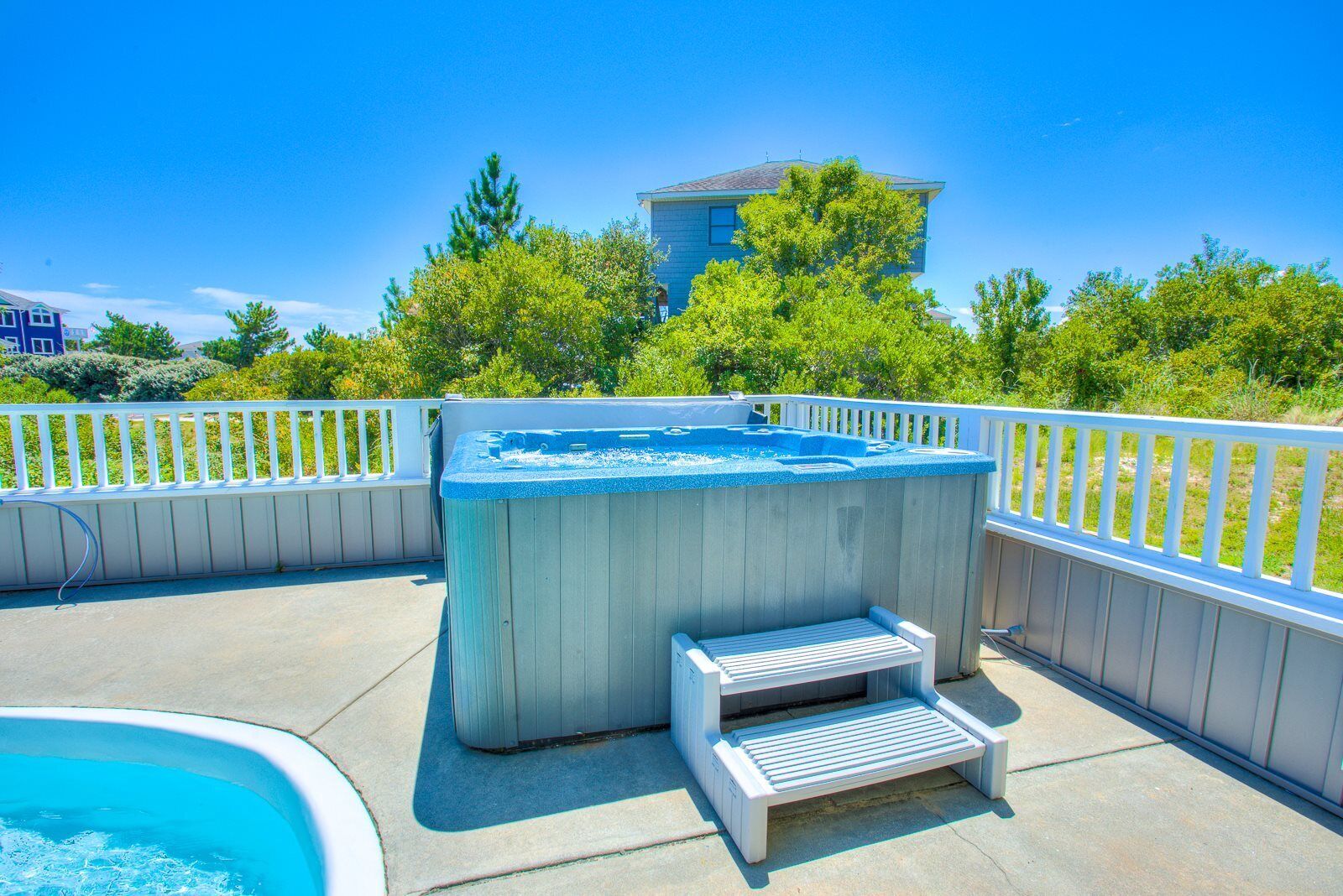 A hot tub on a deck with a pool, surrounded by a white fence and trees.