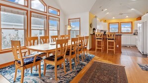 Un comedor amplio con sillas y una mesa de madera, una cocina con armarios blancos y una barra, y ventanas grandes que ofrecen vistas al exterior.