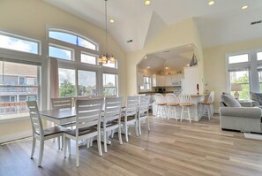 A spacious living room with a dining area, large windows, and a kitchen in the background. - Sea n Stars: Pool, Hot Tub, 5-Min Walk to Beach (Corolla)