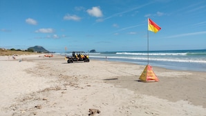 A beach with a lifeguard flag, a lifeguard tower, and a beach buggy. - Omanu Beach Break - Walk across the road to the beach, close to golf (Mount Maunganui)