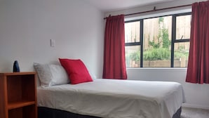 A bedroom with a bed, a wooden nightstand, a window with red curtains, and a view of greenery outside. - Omanu Beach Break - Walk across the road to the beach, close to golf (Mount Maunganui)