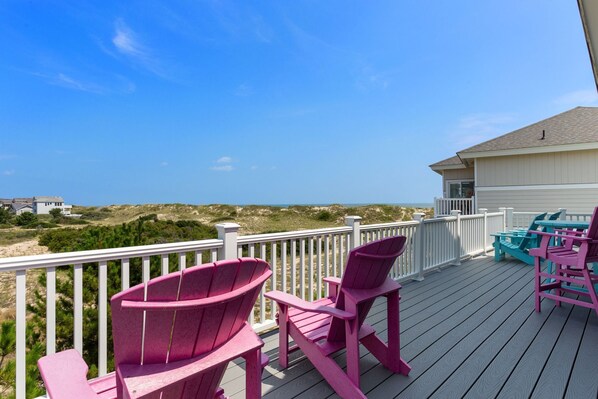 A wooden deck with pink and turquoise Adirondack chairs, overlooking a beach and dunes.