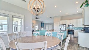 A spacious kitchen with white cabinets, a central island, and a dining area with a round wooden table and white chairs. - Bikinis & Martinis: Walk Beach, Pool, Hot Tub (Kitty Hawk)
