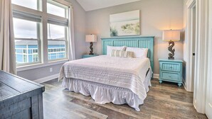 A bedroom with a large bed, two bedside tables, a window with curtains, and a painting on the wall. - Bikinis & Martinis: Walk Beach, Pool, Hot Tub (Kitty Hawk)
