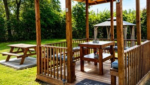 Um gazebo de madeira com mesa de piquenique e bancos, rodeado por um gramado.