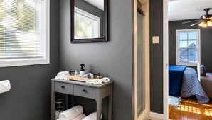 Uma casa de banho moderna com parede escura, toucador com toalhas, espelho e vista para um quarto com cama e ventoinha de teto.