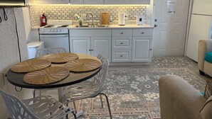Una cocina compacta con armarios blancos, microondas, estufa y lavabo. Hay una mesa de comedor con individuales tejidos y sillas de metal.