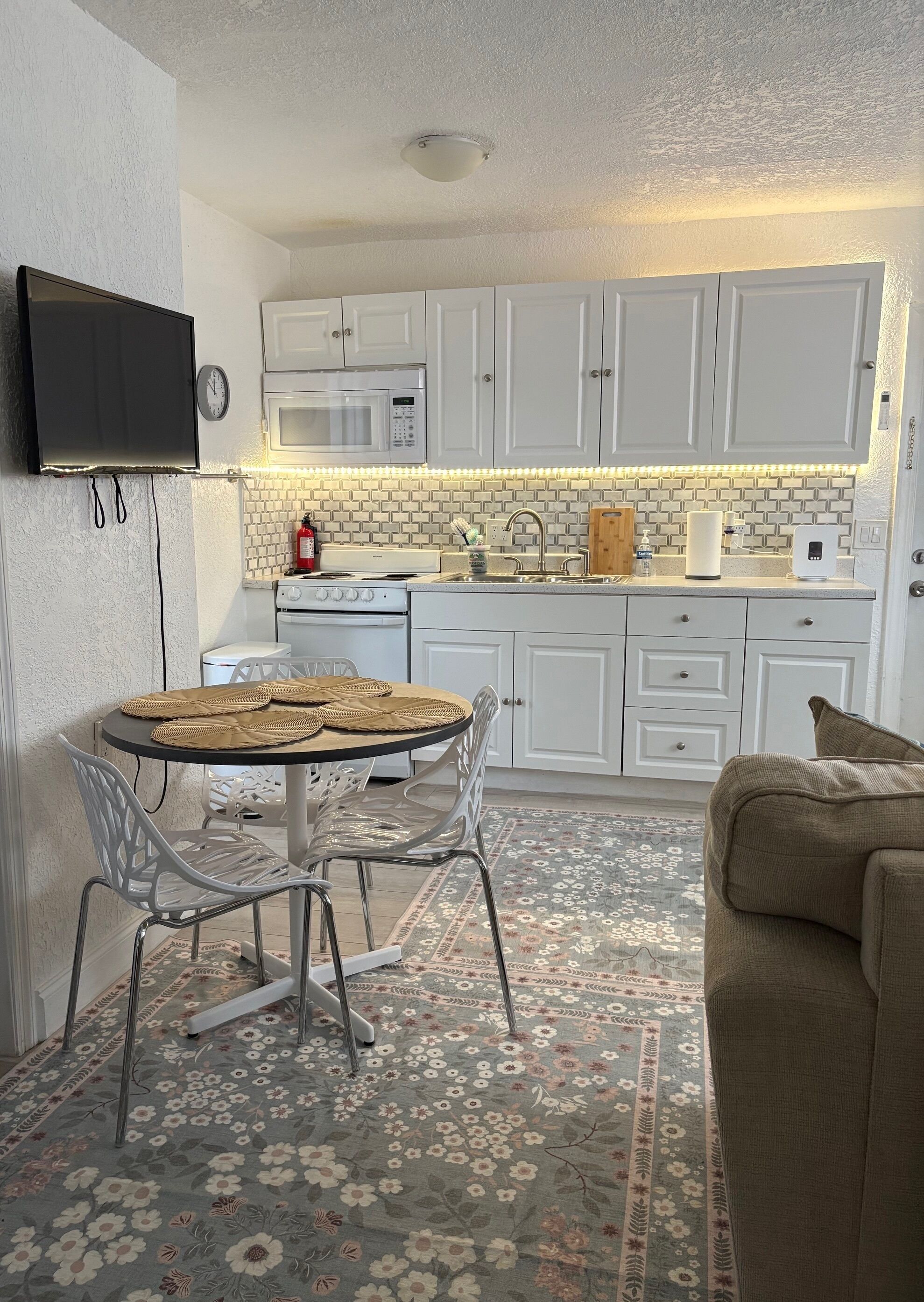 Cocina compacta con gabinetes blancos, microondas, estufa y comedor con mesa redonda y sillas.