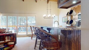 A kitchen with a bar area, wooden chairs, a bookshelf, and a view through the glass doors. - Pet Friendly | Close to Lifts | Spacious | Garage Access | Angel Fire Commons C8 (Angel Fire)