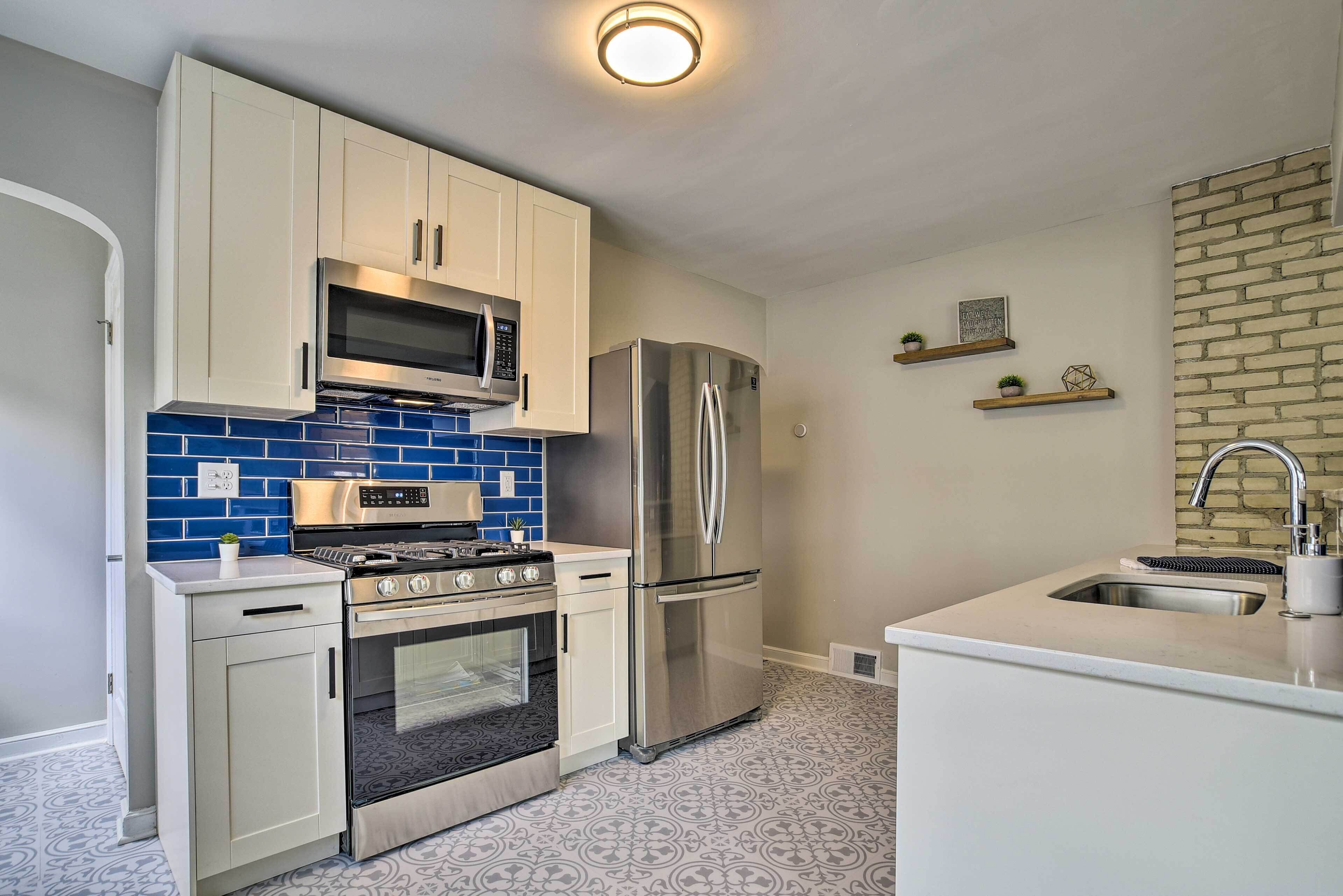 A modern kitchen with white cabinets, a stainless steel refrigerator, a microwave, and a stove. There are blue tiles on the backsplash and a brick wall on one side.