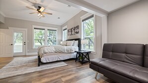 A bedroom with a bed, a brown leather sofa, a ceiling fan, and large windows. - Renovated w/ Pool, Golf, Games & Backyard Activities! (Dripping Springs)