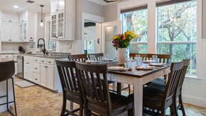 A kitchen with white cabinetry, a dining table set for four, and large windows offering a view of trees. - Renovated w/ Pool, Golf, Games & Backyard Activities! (Dripping Springs)