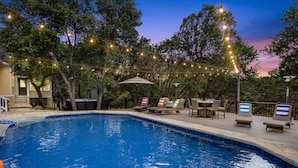 A swimming pool with lounge chairs and umbrellas, surrounded by trees and string lights. - Renovated w/ Pool, Golf, Games & Backyard Activities! (Dripping Springs)