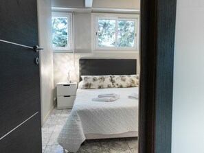 Ein Schlafzimmer mit einem Bett, einem Nachttisch, einer Lampe und einem Fenster mit Blick auf Bäume.