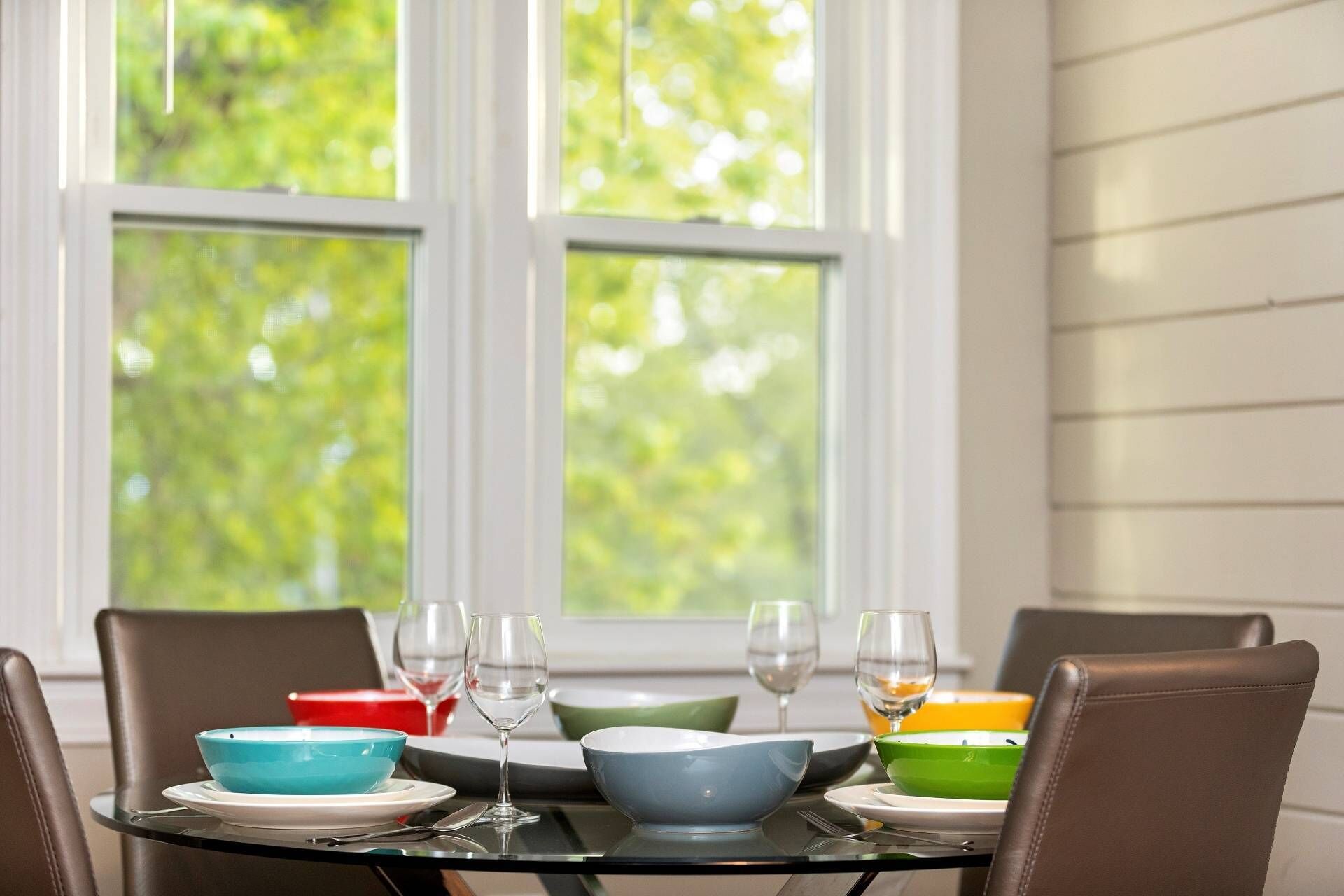 A dining table set with colorful bowls and wine glasses, positioned near a window with a view of trees.