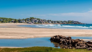 A beach with houses in the background and a rocky outcrop in the foreground.