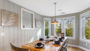 A dining area with a wooden table set for four, surrounded by large windows with a view of trees.