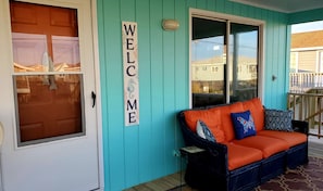 Ein außenstehendes Gebäude in Türkis mit einem 'WELCOME'-Schild, ein Korbsessel mit orangefarbenen Kissen und eine Holzterrasse.
