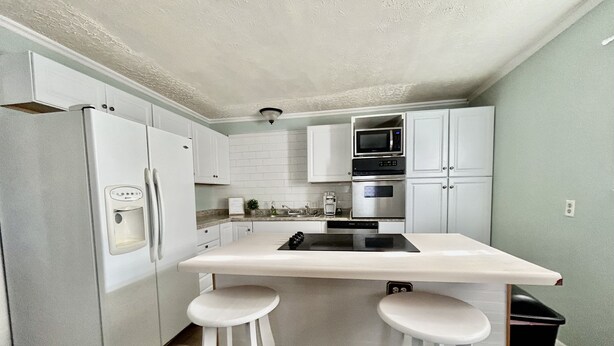 A compact kitchen with white cabinets, a built-in microwave, an oven, a refrigerator, and a small island with stools.