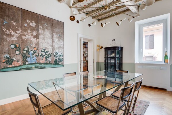 A dining area with a glass table, wicker chairs, a large floral painting, a black cabinet, and a window with shutters.