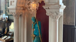 Una estatua de una figura vestida con una túnica, sosteniendo una cruz, sobre un fondo de columnas blancas con un candelabro colgante.