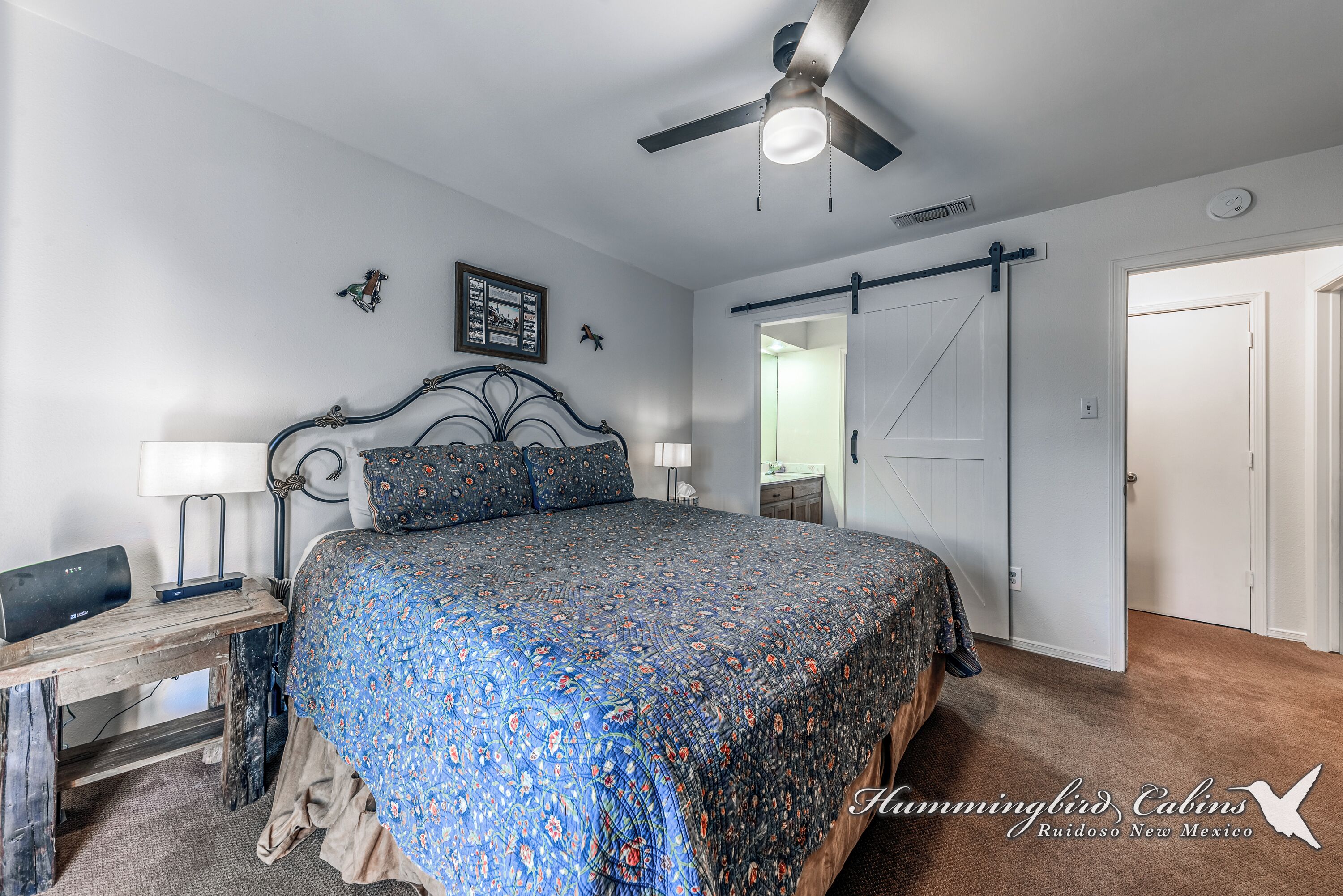 A bedroom with a bed, a ceiling fan, a desk with a lamp, and a door leading to another room.