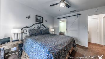 A bedroom with a bed, a ceiling fan, a desk with a lamp, and a door leading to another room.