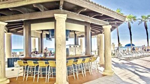 Un bar frente a la playa con mesas al aire libre, vistas al océano y palmeras al fondo.