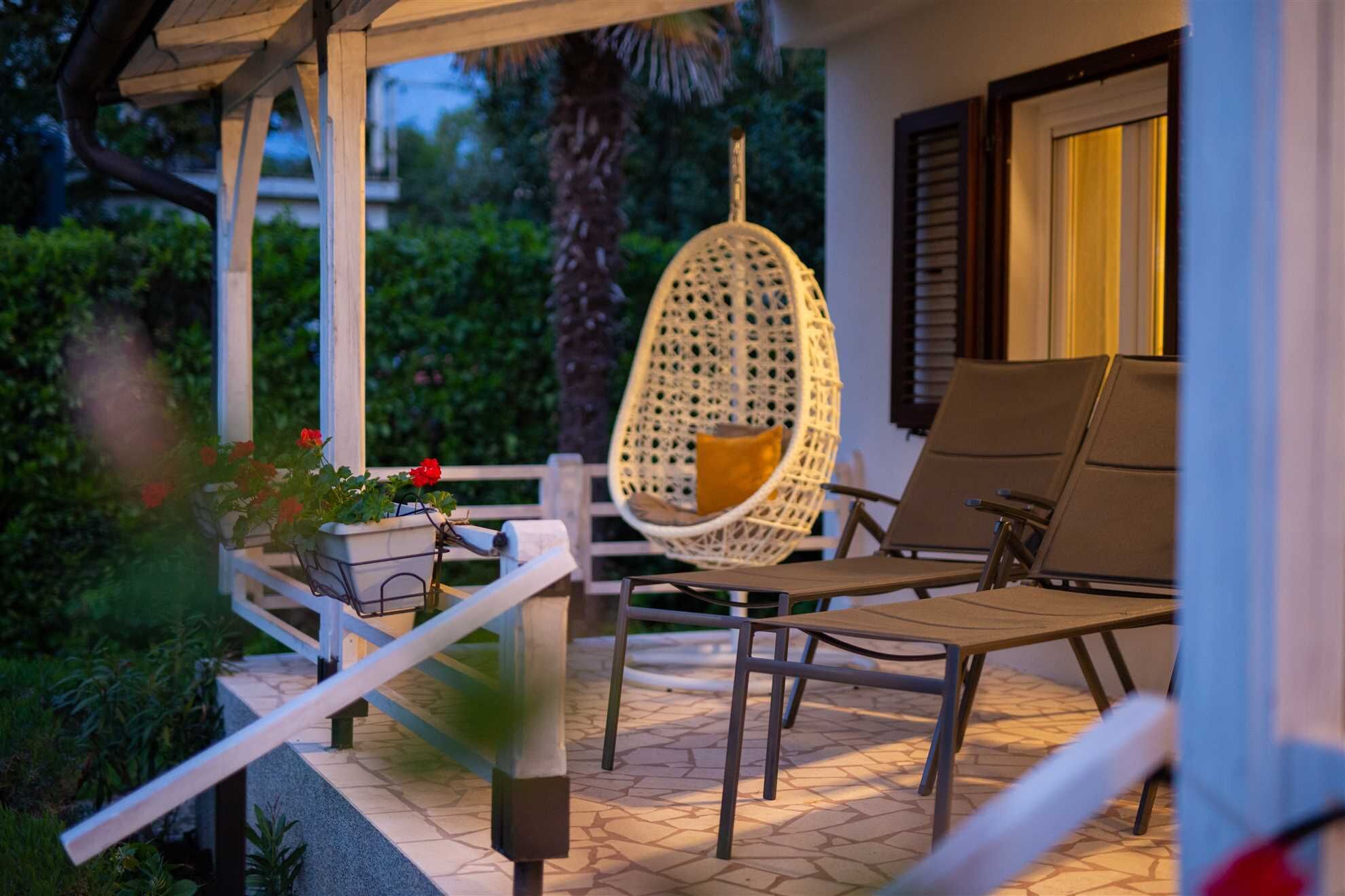 A covered outdoor seating area with a hanging chair and a table, surrounded by greenery.