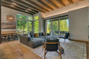 Un salón amplio con un sofá seccional gris grande, una mesa de centro redonda y una silla de mimbre. Varias ventanas ofrecen vistas al exterior.
