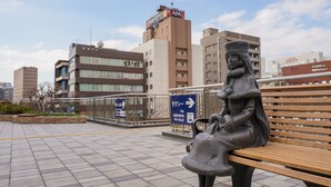 Une statue représentant une femme assise sur un banc, dans un environnement urbain, avec des bâtiments en arrière-plan et un panneau indiquant la direction à suivre pour trouver une station de taxis.