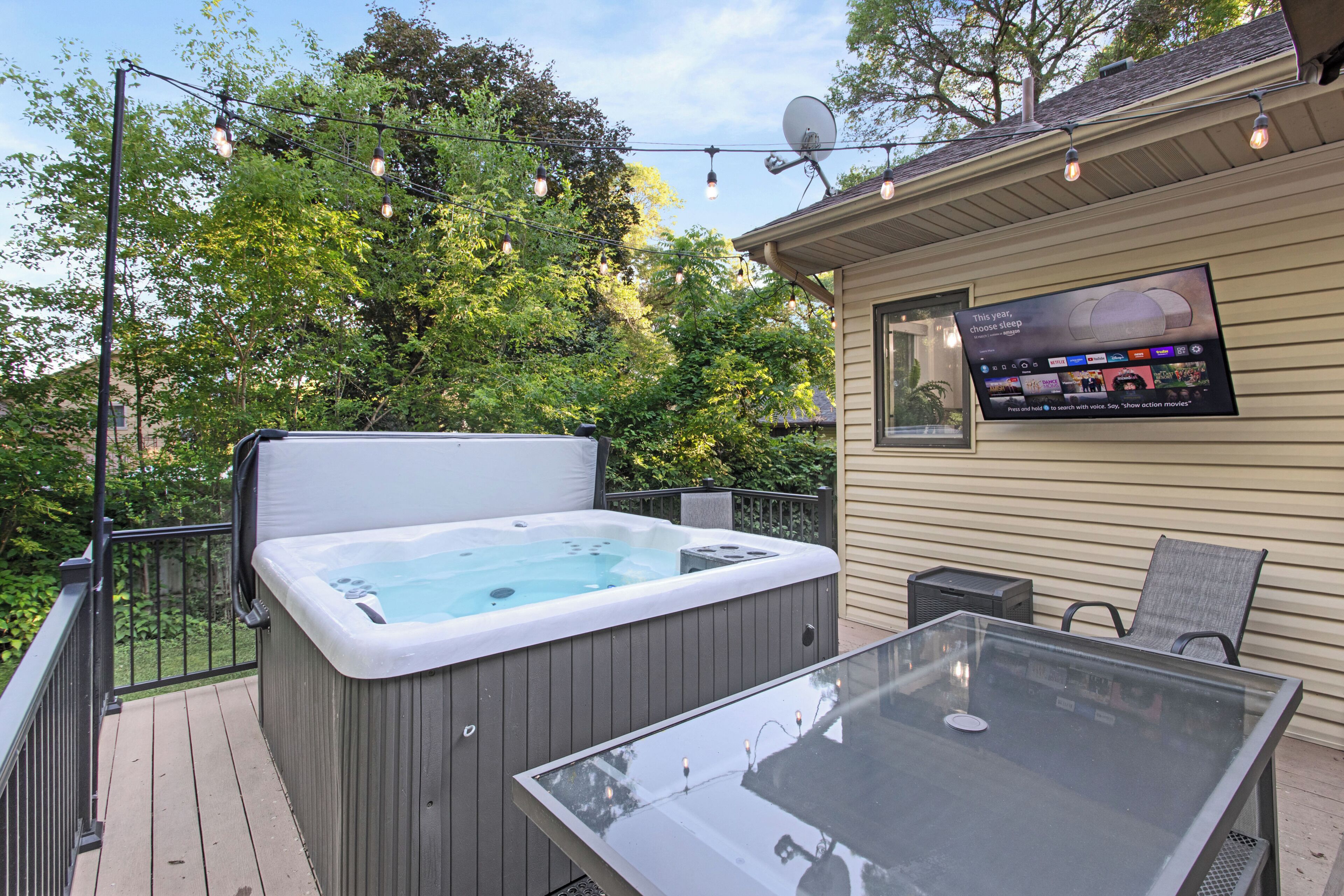 A hot tub on a deck with a glass table and chairs, surrounded by trees and a screened area with a TV.