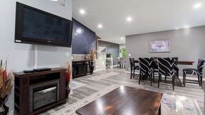 Una sala de estar moderna con un televisor de pantalla plana, un mueble de madera oscura para equipo audiovisual y una zona de comedor con sillas estampadas.