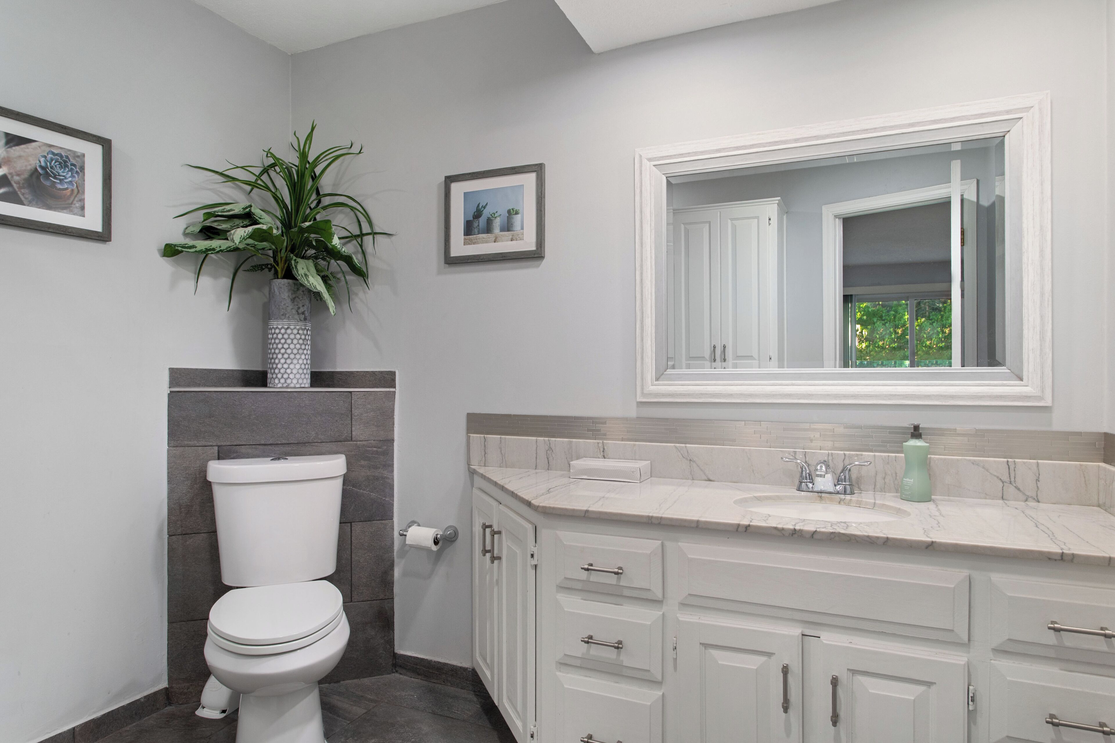 Une salle de bain avec des toilettes, un lavabo avec un miroir au-dessus, une plante en pot et un tableau encadré au mur.