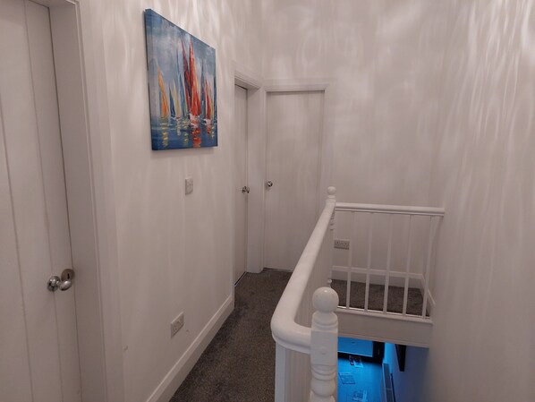 A hallway with white walls, a painting of sailboats, and a white railing with a blue light underneath.
