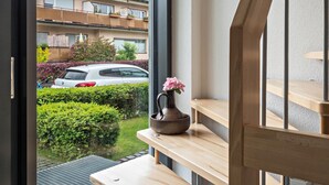 Une vue depuis une fenêtre donnant sur un balcon avec un banc en bois, une plante en pot et une voiture garée à l’extérieur.