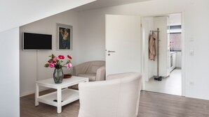 Une pièce moderne et minimaliste, avec un canapé blanc, une petite table sur laquelle est posé un vase de fleurs, un téléviseur à écran plat et une porte donnant sur un coin cuisine.
