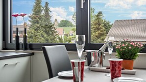 Une table à manger avec un verre à vin, une tasse rouge et une assiette dressée pour deux personnes, donnant sur un vignoble et un bâtiment, le tout visible à travers une fenêtre.