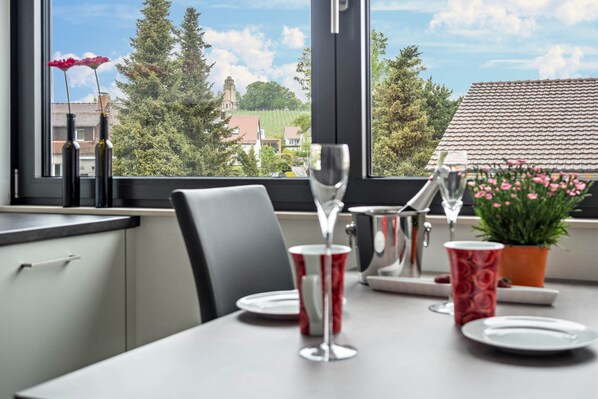 A dining table with a wine glass, a red cup, and a plate set for two, with a view of a vineyard and a building through a window.