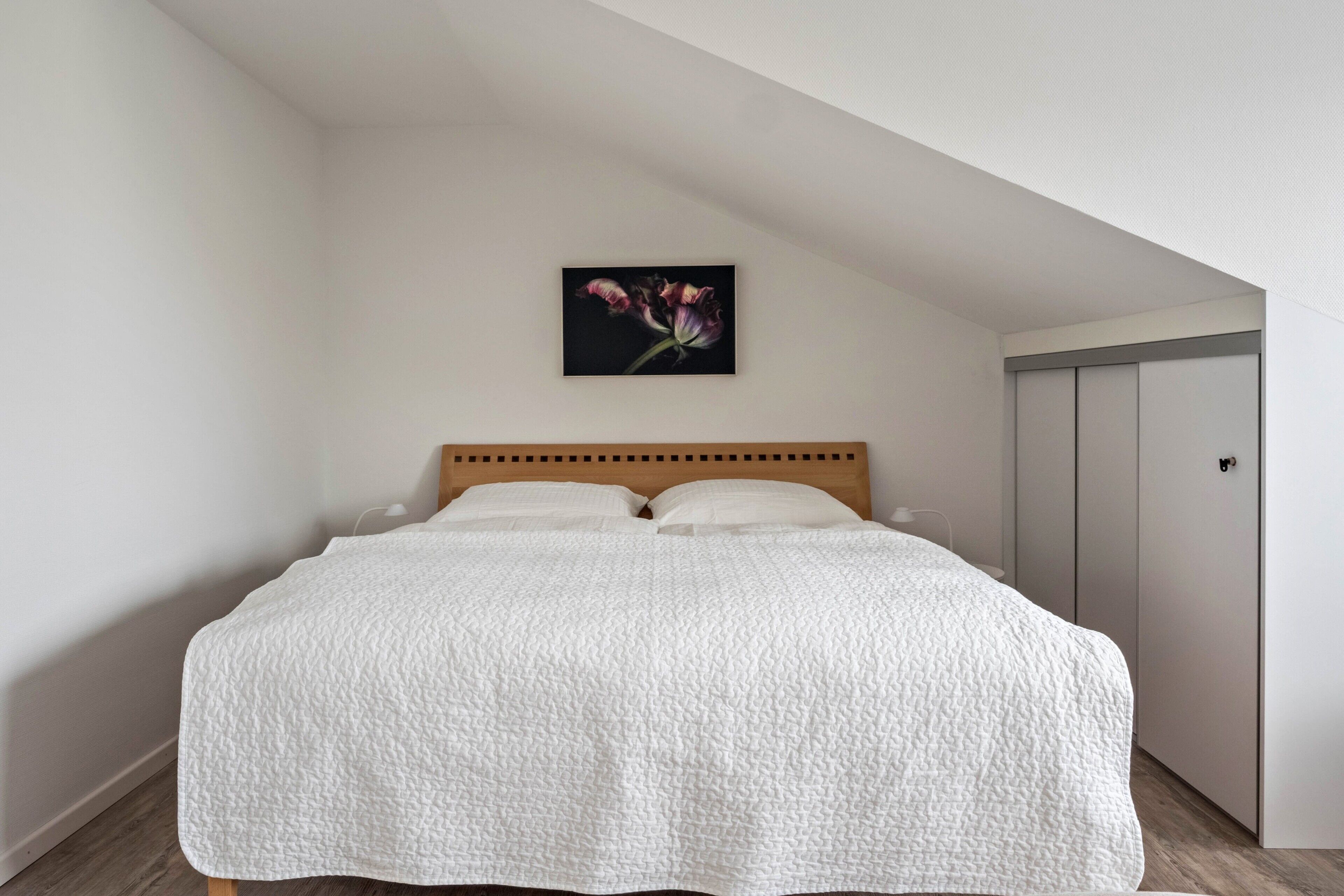 A bedroom with a wooden headboard, a white quilted bedspread, a wall-mounted picture of flowers, and a wardrobe.