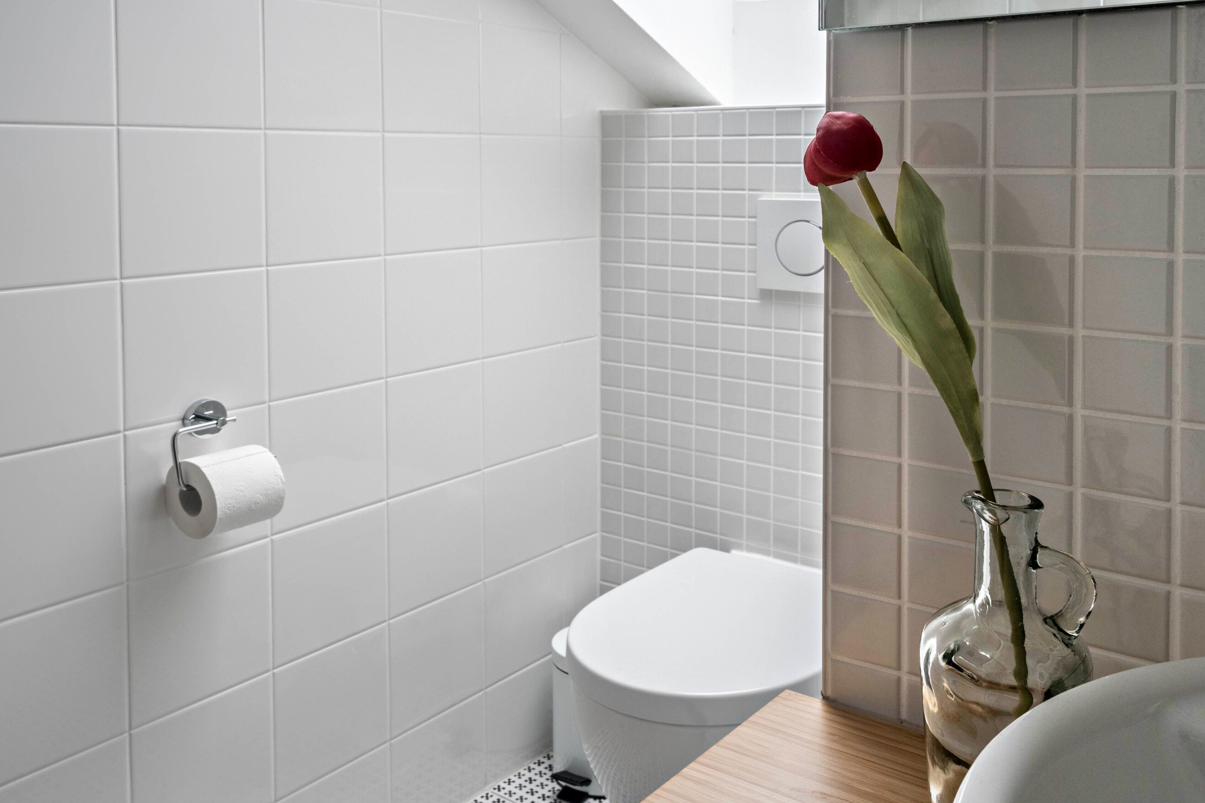 Een badkamer met witte tegels, een toilet, een rol toiletpapier en een vaas met één rode bloem.