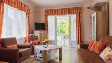 Ein Wohnzimmer mit Sofa, Sessel und Couchtisch. Ein Fernseher steht auf einer Kommode, und es gibt einen Balkon mit Tisch und Stühlen.