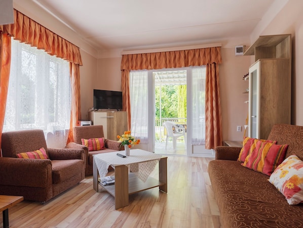 A living room with a sofa, armchair, and a coffee table. There is a TV on a cabinet, and a balcony with a table and chairs. - Gizella by Interhome (Balatonlelle)