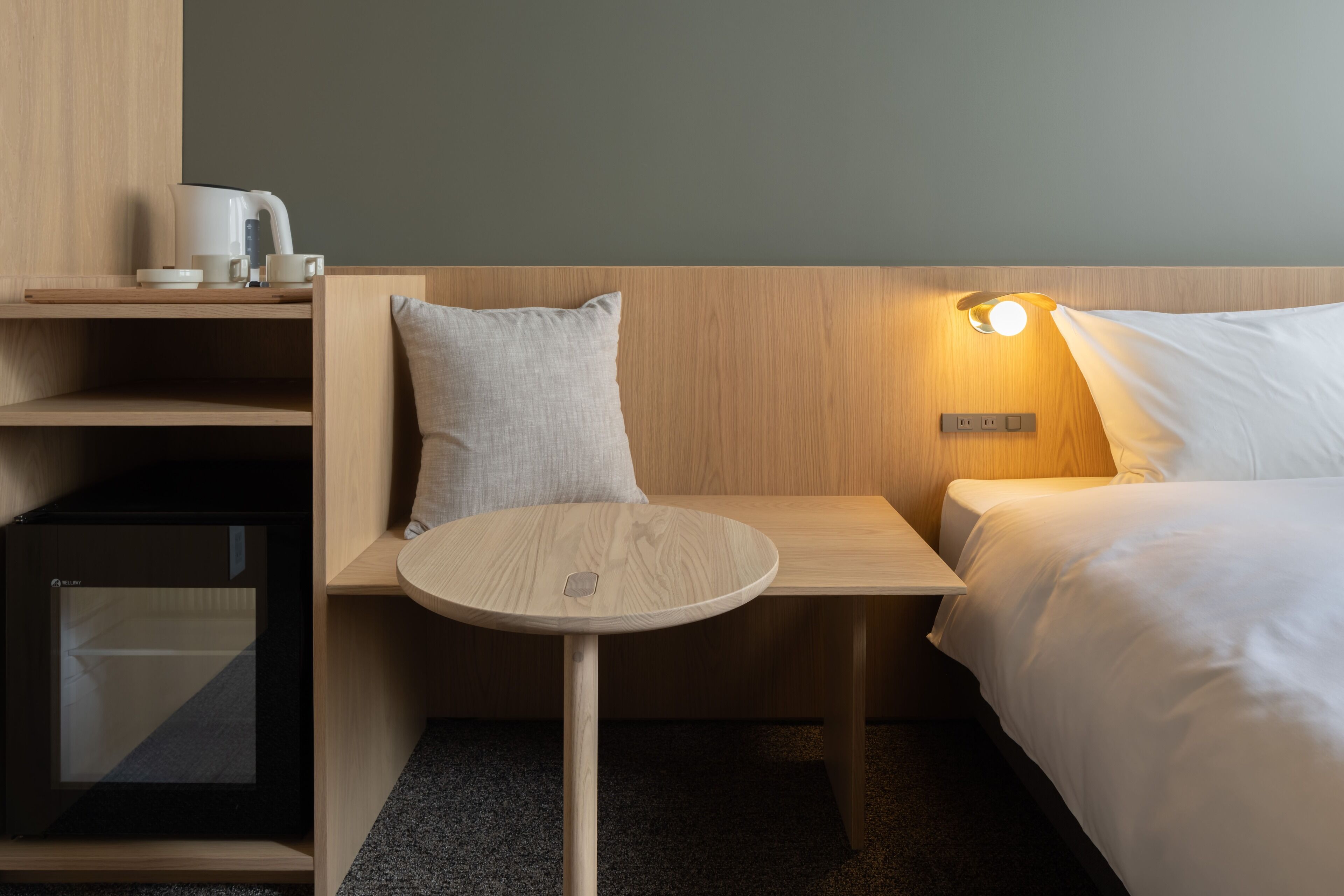 Ein Hotelzimmer mit einem hölzernen Kopfteil, einem Nachttisch mit Lampe, einem Bett mit weißer Bettwäsche und einer kleinen Ablagefläche mit Wasserkocher und Tassen.