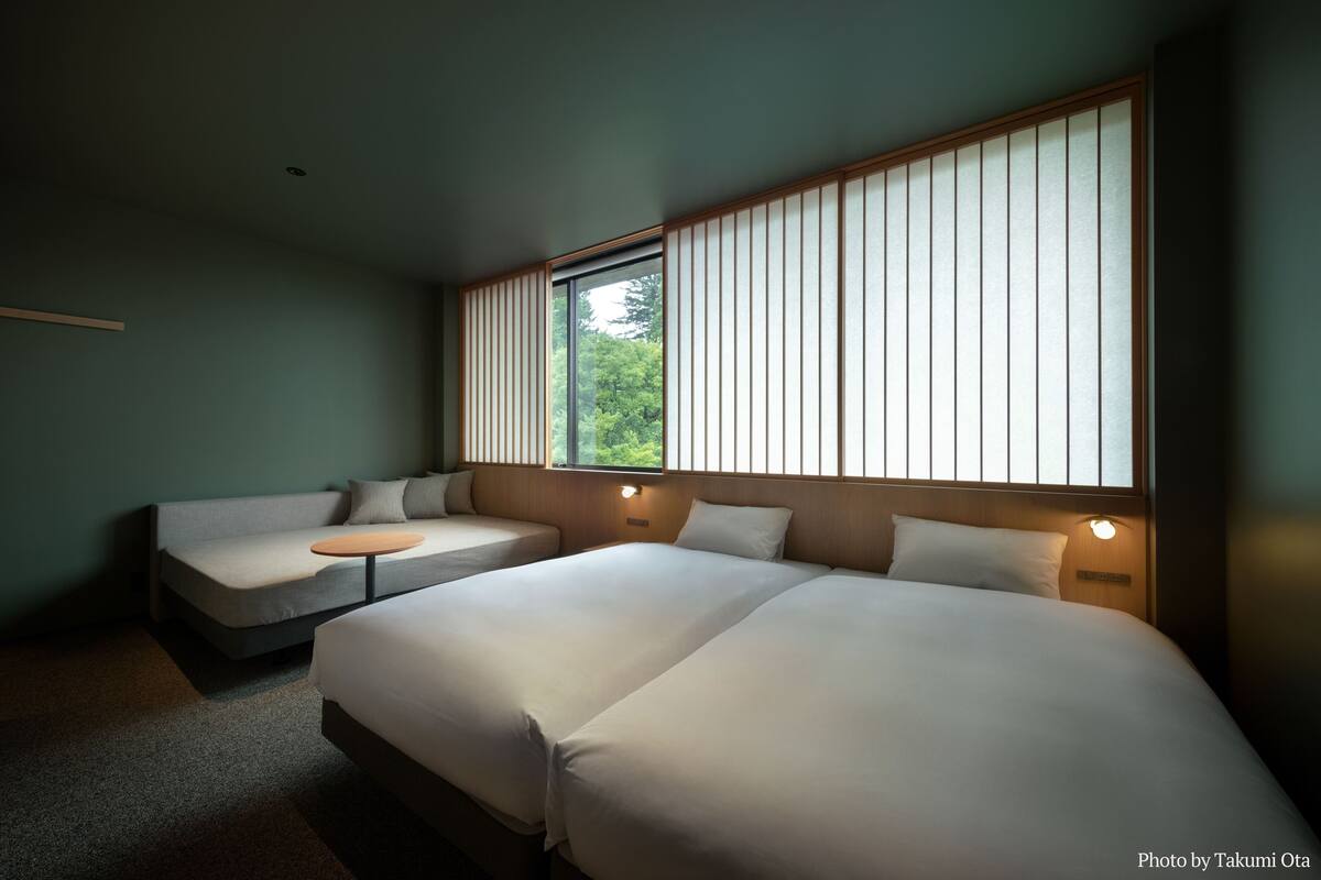 Een moderne hotelkamer met twee grote bedden, een kleine bank en een raam met verticale jaloezieën.