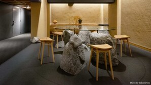Een modern interieur met een glazen tafel, omringd door houten krukken, grote rotsen en een vaas met gedroogde planten.